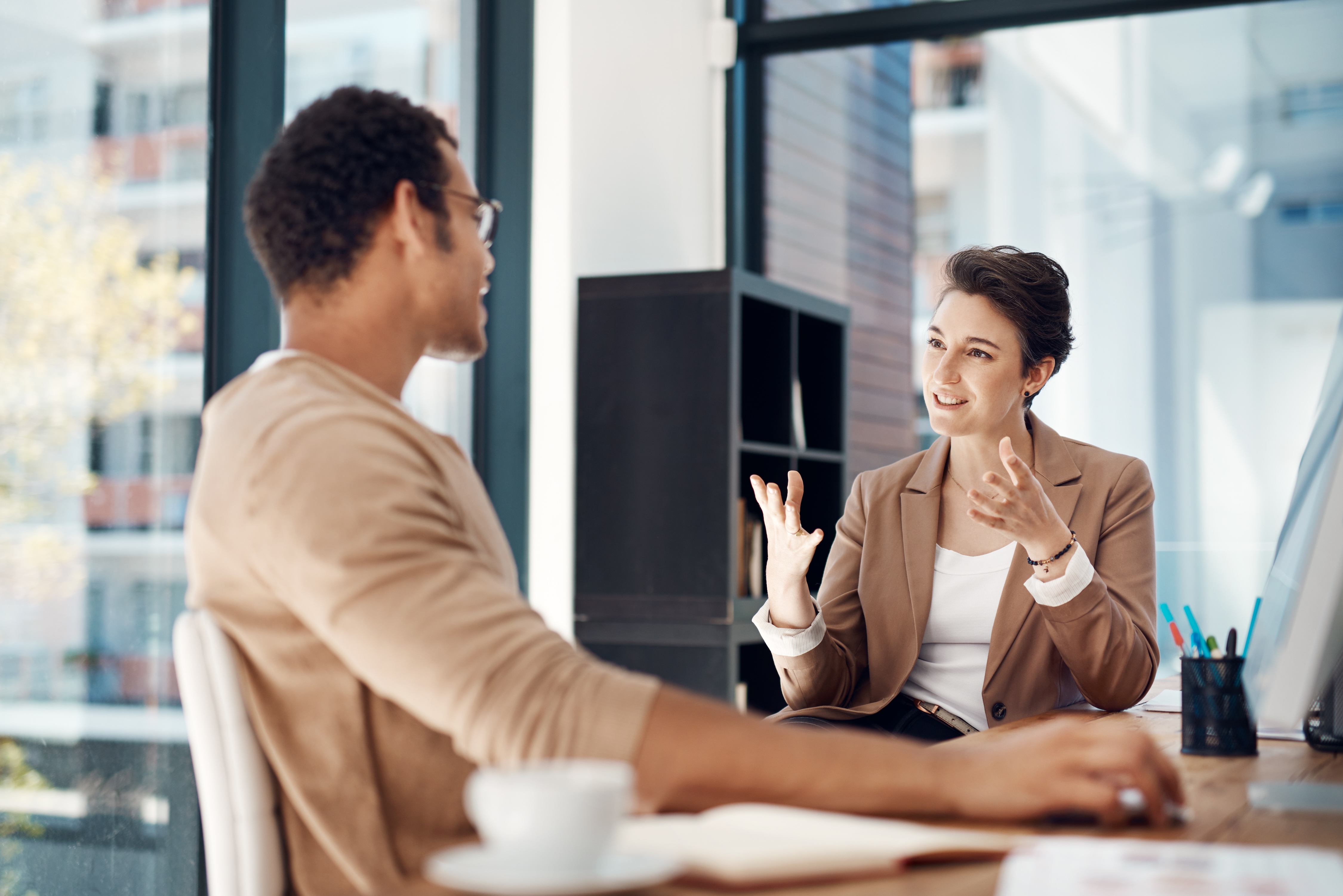 Man and woman converse at an office desk, woman gesturing.